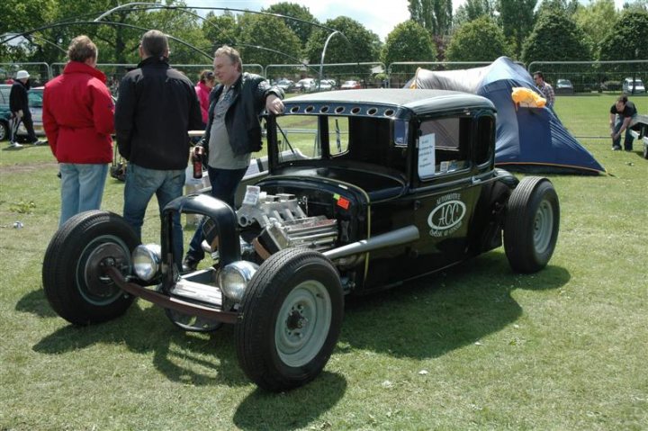 Fins and Chrome - Page 1 - North East - PistonHeads - The image captures a charming scene of a vintage black car parked on a lush green field. A group of people, casually dressed in jeans and t-shirts, are gathered around the car, engaged in lively conversation. The car's classic lines and shiny exterior suggest it is well-maintained, adding a sense of nostalgia to the scene. One of the individuals leans nonchalantly against the hood of the car, their body language relaxed and at ease. In the background, a tent is pitched, suggesting an outdoor event or gathering. The sky is a clear blue, indicating a bright, sunny day.