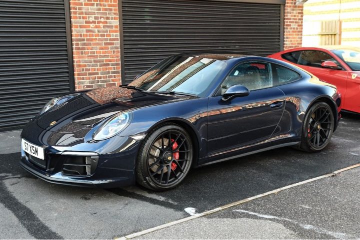 991 GTS Must haves - Page 176 - 911/Carrera GT - PistonHeads - The image presents a scene of two sports cars in a parking lot. The most prominently featured is a black Porsche sports car, positioned on the left side of the frame, facing towards the right. The Porsche's sleek design and glossy black finish suggest it's a high-end model. To the right of the Porsche, there is a red sports car, which is smaller in comparison, capturing less attention due to its placement in the frame. Both cars are parked on asphalt, and a closed garage door is visible in the background, adding context to the location. The focus of the image is clearly on the Porsche, highlighting its status as a luxury vehicle.