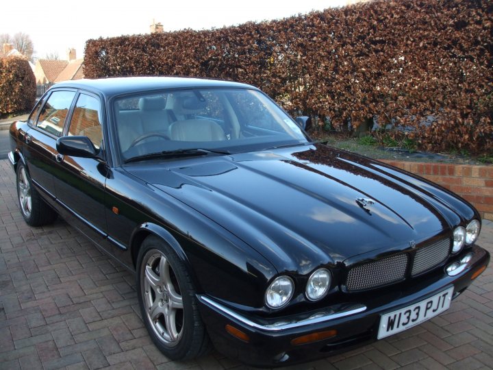 To XJR or not to XJR - Page 1 - Jaguar - PistonHeads - The image shows a black luxury car positioned on what appears to be a brick driveway. The car's sleek design is accentuated by shiny, well-maintained paint, and the front grille features distinct badges, indicative of the vehicle's high-end status. The vehicle's open doors are facing the viewer, revealing the well-crafted leather interior and the sunroof. Surrounding the driveway is a neatly trimmed hedge, which provides a serene backdrop to the car. The overall condition of the vehicle and its surroundings suggest a well-kept and cherished possession.