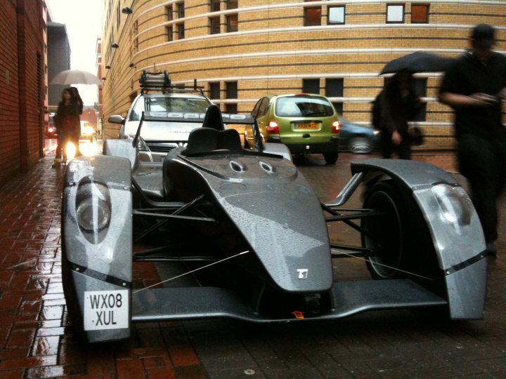 Birmingham Pistonheads Forgot Icc Lovely Beast - The image captures a dynamic cityscape on a rainy day. Dominating the scene is an open-top, silver-and-black concept race car, which is parked on the sidewalk to the left of the frame. The car is sleek and mechanical, reflecting the wet bricks that make up the sidewalk. 

In the background, a pedestrian is walking with an umbrella nearby, adding to the urban atmosphere. The sidewalk itself is wet, reflecting the ambient lighting and adding a sense of atmospheric depth to the image. The buildings lining the street appear modern, suggesting a contemporary urban setting. The overall image conveys a sense of motion and dynamic energy.