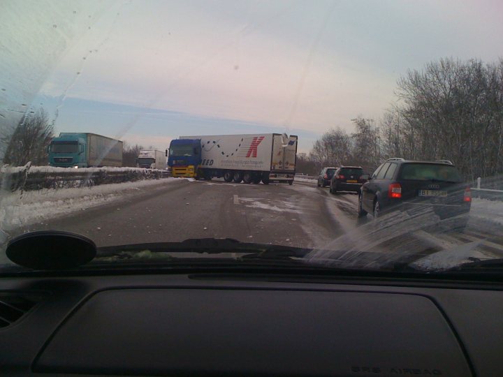 Pistonheads - The image shows a multi-lane highway during adverse weather conditions. There's a large truck in one lane that appears to have experienced an accident or is suffering a mechanical issue, as it is stranded on the side of the road. The truck takes up two lanes. In the other lanes, there are various cars, including at least one sports car. There's a clear buildup of snow and slush on the road and along the roadside. The weather elements, highlighted by a streak across the sky, suggest it might be snowing at the time. The cars, including the one we see through the windshield of a car in the foreground, are in traffic due to the disruption caused by the stranded truck.