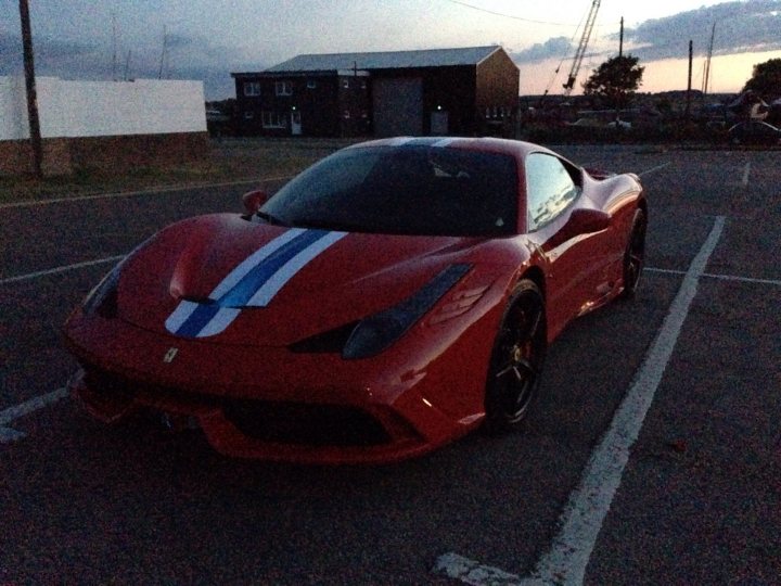 New wheels.. Something speciale.. - Page 1 - East Anglia - PistonHeads - This image captures a scene at sunset. The main focus is a sleek, red sports car parked in a parking lot. The car is adorned with a blue and white stripe running down its length. The setting sun casts a warm glow on the car, enhancing its vibrant, red hue. In the background, there is a white building with a gray roof. The sky is transitioning from the warm glow of the sunset to a darker blue, signaling the arrival of evening.