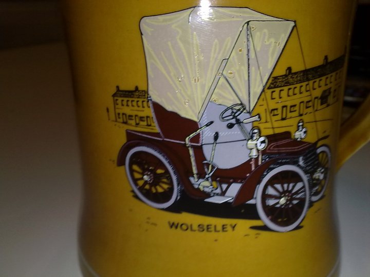 Century Pistonheads Turn Wolesley - The image features a large, ornate mug with a yellow body and a handle on the left side. The mug is adorned with an illustrated scene depicting a vintage brown car with a canopy, pulled by two white horses. The name "Wolseley" is visible, suggesting the model of the depicted car, and below the illustration is another text that reads "Wolseley," indicating the brand. The mug is placed on a surface, and to the right of it, another, smaller mug can be partially seen.