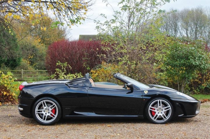 A black and white photo of a car parked in a field - Pistonheads - The image depicts a black convertible sports car parked on a gravel surface amidst autumn foliage. The car has a sleek design with prominent red brake calipers. In the background, there is a landscape of trees and plants that suggest a natural, possibly rural setting. The sky is overcast, providing a neutral backdrop for the vivid colors of the leaves and the car itself.