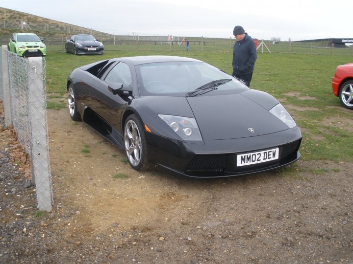 Thank you. - Page 1 - East Anglia - PistonHeads - This image features a parking lot with a lot of cars. The most distinctive car is a sleek, black sports car with a convertible top. It has a low stance and sporty wheels. Not far from the sports car, there are two other cars parked. The background shows a fence and a hill. It appears to be a bright, cloudy day. The license plate of the sports car is partially visible and reads "MMM Q2 DEW".