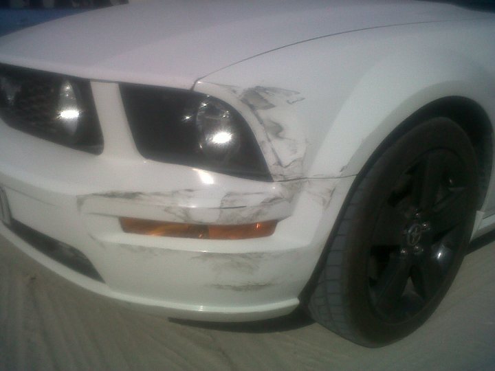 Pistonheads - The image shows the side of a white car with noticeable damage. The car appears to be a muscle car, judging by the distinctive front grille. A large scrape covers the front fender, indicative of a recent accident. The vehicle is in motion, as suggested by the blurred background and ground in the distance. The tire on this side of the car is a standard tire without any visible spikes or additional modifications.