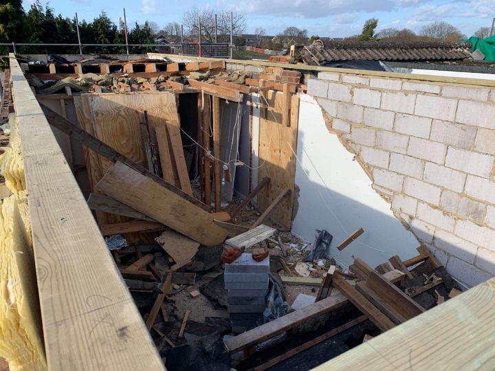 Build thread - bungalow to house - Page 2 - Homes, Gardens and DIY - PistonHeads - The image shows a partially demolished building. The structure is incomplete, with exposed wooden studs and beams visible through the open roofing. The ground floor is intact, but the upper levels are missing or damaged. Debris is scattered throughout the space, suggesting recent work or destruction. There's a hole in the wall on the left side of the building, revealing an unfinished interior with exposed wiring. In the background, there are trees and some structures that appear to be intact buildings. The scene suggests a site undergoing construction or renovation.