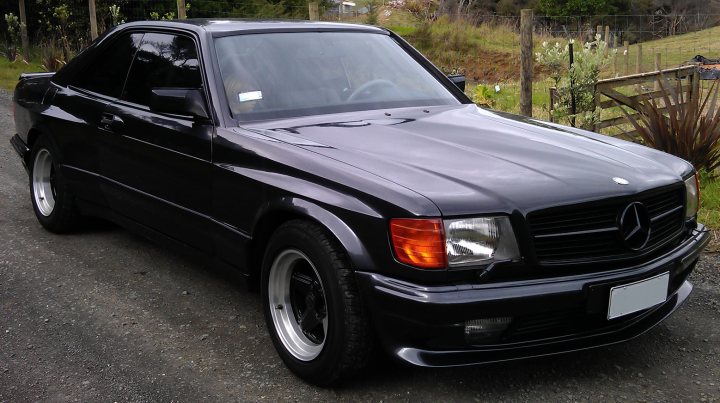 A long shot, but anyone know what happened to this SEC?  - Page 1 - Mercedes - PistonHeads - The image portrays a black Mercedes-Benz car, parked on a gravel drive. The car is facing towards the right side of the image, with a noticeable light glare on the bonnet, suggesting recent use or the presence of the sun. The license plate is visible at the front of the car. The car's sleek design and the sunlight give it a shiny appearance. It appears to be a still day, and the car's position suggests it is parked and not currently in motion. The presence of grass and partial fence in the background indicates a rural or semi-rural setting.