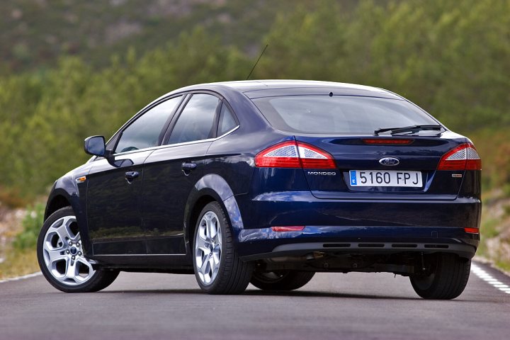 Pistonheads Facelifts Hell - The image features a dark blue four-door sedan on a road with a green forested background. The car is captured in motion, driving on the right side of the road. Its modern design is characterized by distinctive headlights, a prominent grille with the brand's logo, and five-spoke alloy wheels. The license plate is visible and reads "5160 FPJ," indicating the car is likely from the European registration system.