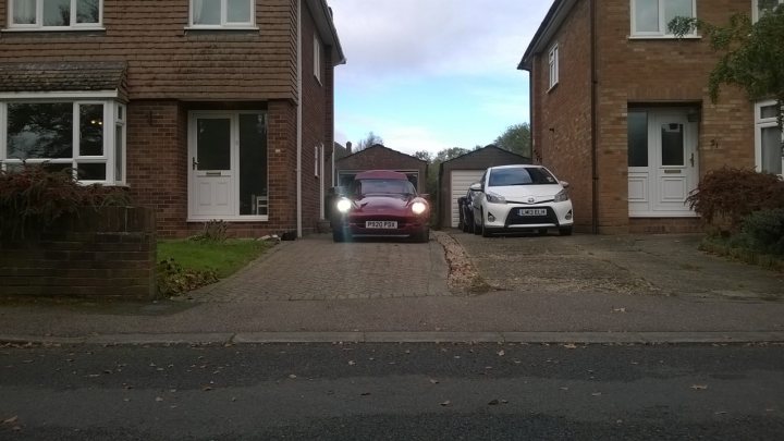 LED Headlights - Page 1 - Chimaera - PistonHeads - In this image, a neatly organized residential street is lined with terraced houses and a blue van. The vans are parked in driveways between the houses, which predominantly feature brick exteriors with white doorframes. A small grassy area borders the road on one side. The sky overhead is partially cloudy, suggesting a calm, overcast day. A few objects, including a potted plant and a sign, add to the residential charm of the scene.