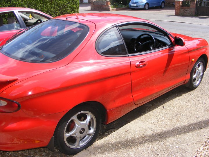 Pistonheads Coupeshed - The image showcases a vibrant red sports car parked on a gravel driveway. The car, which is sleek and shiny, features a glossy finish that reflects the surrounding environment. It has a sloping roofline and a spoiler, adding to its sporty design. The vehicle appears to be new or well-maintained, suggesting careful ownership. It is surrounded by plants, adding a touch of nature to the scene. The car is the central focus of the image, with its bright color and polished exterior drawing the viewer's attention.