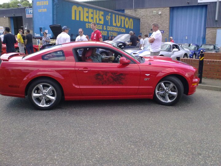 Sat Pistonheads Sports Atlantic July Bbq - This image captures a scene at the "Meek's of Luton" storage facility. The main subject is a bright red sports car parked in front of the building. The car's visor and wheel rims have graffiti on them, adding a sense of urban grit to the scene. In the foreground, there are several people standing, possibly observing the car or waiting in line. In the background, other cars are visible, hinting at the storage facility's function. The overall color scheme of the image is red, white, and black, with the red of the car standing out against the contrasting blue of the storage facility.