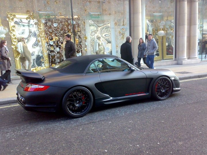 Wheel Options Pistonheads - In the image, a sleek black sports car is parked on the side of the street. The car is positioned diagonally, highlighting its long and low design. The car's contrasting black and red wheels add to its sporty appeal. Several people are standing near the car, engaged in various activities. One person is interacting with the car, possibly opening the door or operating one of the car's features. The overall scene suggests a day in an urban setting.