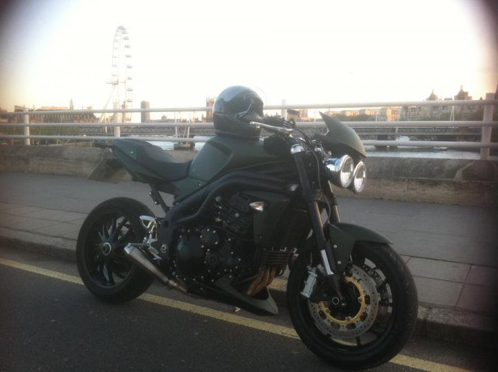 A motorcycle parked on the side of the road - Pistonheads - The image displays a sleek, green motorcycle parked on a street next to a yellow alley line. The motorcycle is facing towards the left side of the image and occupies a significant portion of the view. In the background, there is a round ferris wheel and an urban skyline featuring various buildings. It gives the impression of a modern waterfront city. The photo borders at the bottom, framing the colorful scene within its boundaries.