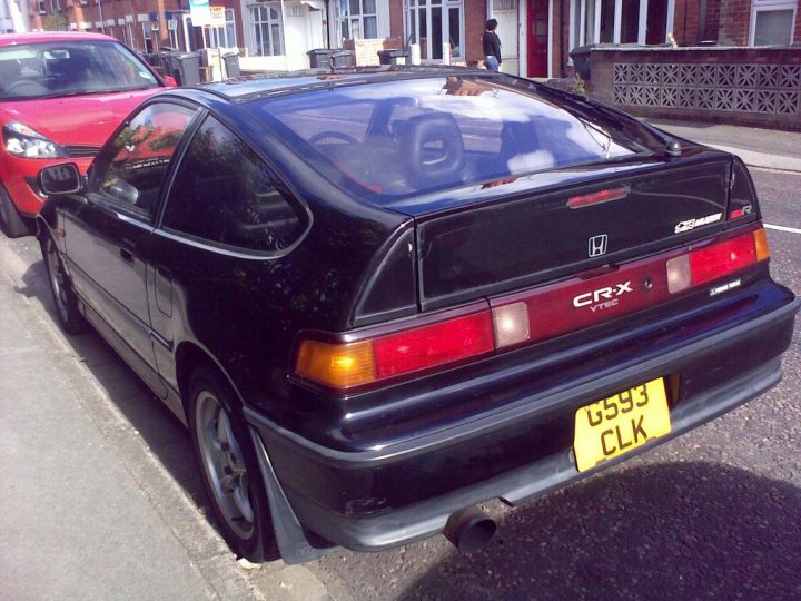 Member Sir Crx Pistonheads - The image shows a small hatchback car parked on a street adjacent to a sidewalk. The car has a black body and a maroon rear. The license plate is yellow with red and black lettering, and the registration reads "GS593 CLK." The rear bumper also has a red reflector and two orange illumination turn signals. Above the license plate, there's a silver emblem with the name "CRX" and below the name, "VTEC" is visible, indicating the engine model. The exhaust pipe of the car is located behind and below the license plate. The street appears to be quiet and there is a red vehicle parked further down the street. The weather seems overcast, as suggested by the lack of bright light in the image.