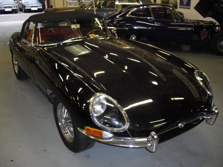 Workshop Tour Pistonheads - The image captures a scene in a garage where two classic black convertibles are parked. The central car, with a striking headlight design, is an older model that appears well-maintained. The car behind it, while not in focus, adds to the vintage atmosphere of the setting. Some of the garage's equipment is visible, including a variety of tools and car jacks. The image evokes a sense of nostalgia and appreciation for automotive history.