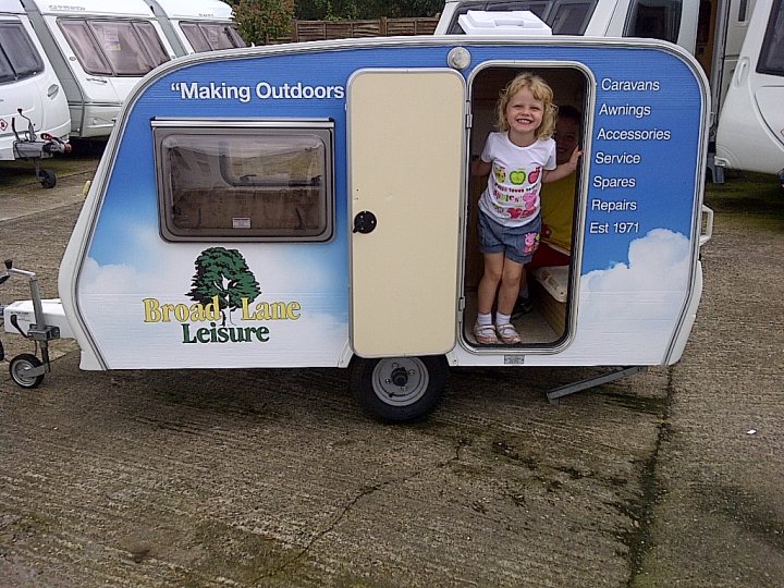 Caravan recommendations with a specific layout - Page 1 - Tents, Caravans & Motorhomes - PistonHeads - In the image, a young girl is joyfully standing in the doorway of a camper, which is uniquely converted into a "Broad Lane Leisure Caravan". The camper is parked on a paved area, possibly a sale yard, as evidenced by several other caravans in the background. The girl is dressed in a white t-shirt and blue jeans, smiling towards the camera, capturing a moment of anticipation and excitement associated with a road trip or vacation. The camper's picture suggests it's a model on display, possibly ready for an outdoor adventure.