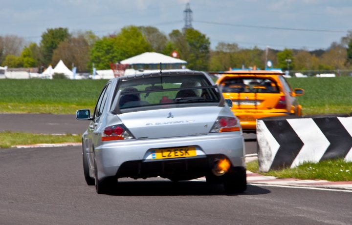 Your Best Trackday Action Photo Please - Page 61 - Track Days - PistonHeads - In the center of the image, a sports car with a black license plate is captured in motion on a red race course, leaving a cloud of dust by its wheels. A phallic emblem on the grille of the car adds a provocative touch. In the background, another car, this time yellow, is visible on the track. Beyond the track, a white tent and other structures hint at a larger event or festival.