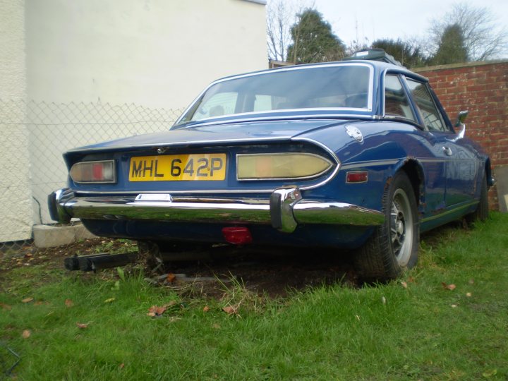 Scrap the Stagg, or give it a new home? - Page 1 - Triumph - PistonHeads - The image depicts a classic blue sedan parked on a grassy area. The car is an older model with distinctive design features. Its license plate is clearly visible, and the vehicle is equipped with large, round headlights. The background includes a fence and a building.