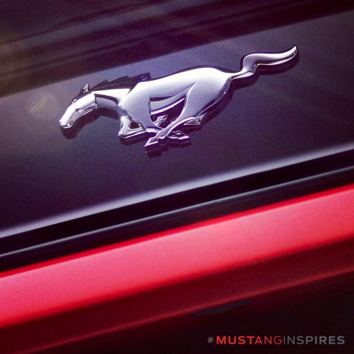 15 MY Mustang picture leaked? - Page 1 - Mustangs - PistonHeads - The image features a close-up view of the emblem of a horse on a car's trunk. The horse emblem is metallic silver, standing out against the dark car hue. The horse is depicted in a stylized pose with its forelegs raised and its tail curved upward, suggesting motion. The car edge, painted in a vibrant red, adds a striking contrast to the emblem, highlighting the logo on the trunk. The background is blurred, with what appears to be a hint of the sky and clouds, suggesting the photograph might have been taken just before sunset or during a cloudy day.