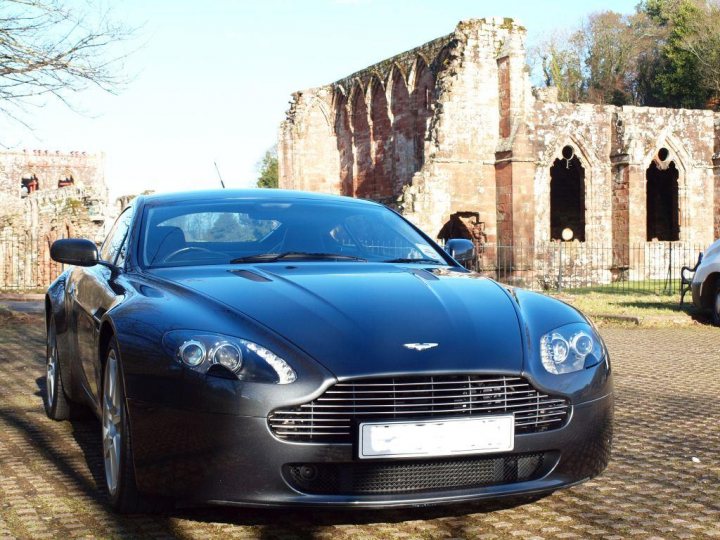 Another New owner - Page 1 - Aston Martin - PistonHeads - The image features a sleek black Jaguar car parked on a gravel driveway. The car is positioned in front of a crumbling building with a large archway. The sky appears clear and blue, suggesting it could be a sunny day. Another car is parked in the background, and there's a glimpse of a green lawn with a tree. The overall setting gives a sense of tranquility and age, possibly at an old or historic location.