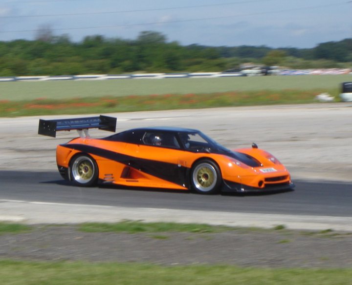 Pistonheads - The image captures a dynamic scene on a race track. Dominating the center of the frame is an orange and black sportscar, driving at a high speed around a bend. The car has a large wing mounted on its back, which contrasts with its smaller wing mounted on its front. These wings are designed for aerodynamic downforce, a technique used in racing to increase vehicle adhesion and maintain stability during high-speed cornering. The background reveals a well-maintained track with a clear sky above, indicating favorable weather conditions for racing.