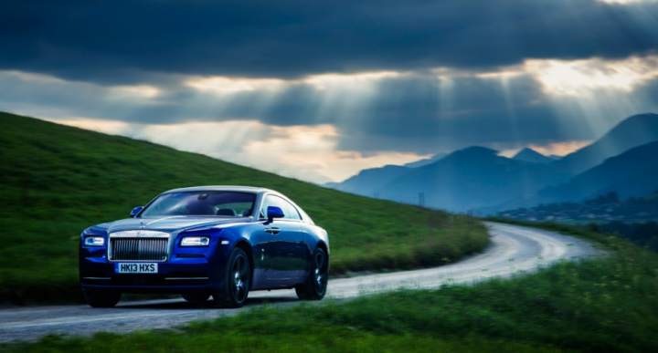 Spotted - Page 1 - Rolls Royce & Bentley - PistonHeads - In the image, a blue luxury car is driving down a winding, country road. The car is large and sleek, positioned in the center of the image, creating a sense of motion. The road is flanked by lush green hills and a grassy verge, giving the scene a pastoral charm. The sky above is cloudy, suggesting a late hour setting. Respectively, the car bears the license plate "HK31 HUO".