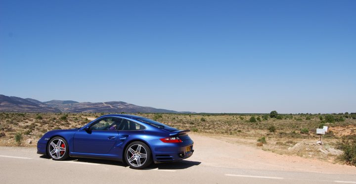 Turbo Spain Vacation Porsche Pistonheads - The image depicts a hard-surface road with a striking blue Porsche parked in the middle. The car is facing to the right, as if it's parked facing away from a hill or a view of mountains. The setting appears to be a desert or arid landscape with sandy ground on both sides of the road. The sky is clear and blue with no clouds visible, enhancing the sense of isolation and emptiness.