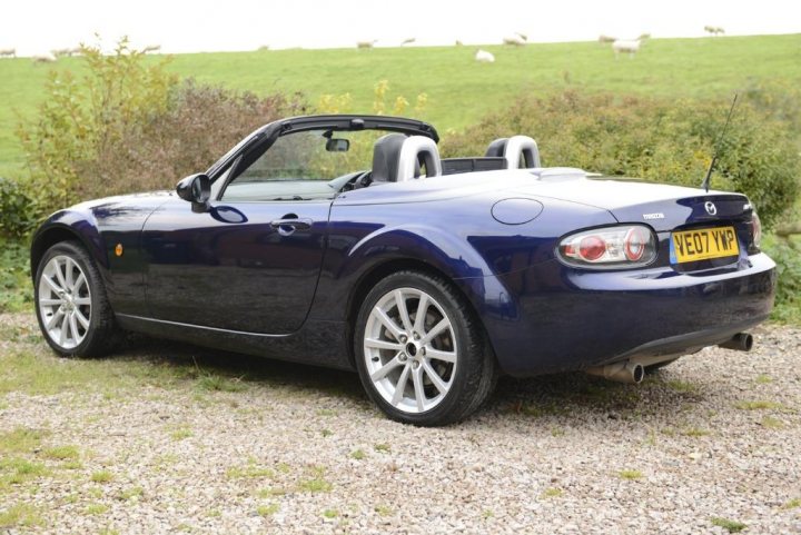 MK3 MX-5 for a 20 year old? - Page 1 - Mazda MX5/Eunos/Miata - PistonHeads - The image showcases a vibrant blue convertible sports car parked on a gravel driveway. Off to the side of the driveway are white sheep grazing, adding a rural charm to the scene. The car's top is down, affording an unobstructed view of its black seats and striped, red and white, license plate. In the background, lush green grass and scattered bushes enhance the natural setting. The picture presents a harmonious blend of interior design and rural tranquility.