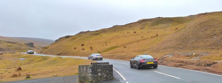 Vanquish II Volante - Page 4 - Aston Martin - PistonHeads - The image captures a winding mountain road, with two cars in motion. The road appears to be in good condition, with clear demarcation for lanes. The mountains in the background are characterized by a rust-like color and are covered with vegetation. The overall setting suggests a remote or rural location, possibly in a region with arid landscapes. The perspective of the image is from a slightly elevated viewpoint, looking down the road and towards the mountains.