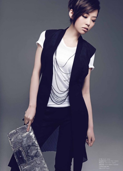 The image features a young woman in a studio portrait. She has short hair and is looking down to her right. She is wearing a blazer with a white t-shirt underneath and black pants. The blazer has a distinctive pattern, and she is holding a metallic clutch in her right hand. Her pose is casual yet stylish, with one hand resting on her hip and the other touching the base of the clutch. The background is a neutral gradient, which contrasts with her attire and accessory.