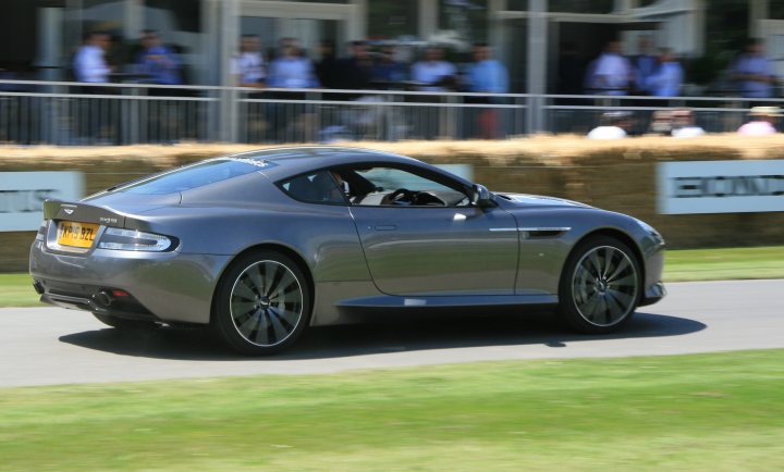 Goodwood moving motor show photos - Page 1 - Goodwood Events - PistonHeads - The image showcases a modern, grey-colored sports car in motion on a racing track. The car's design suggests a high-performance capability, with sleek lines and large alloy wheels. The image is taken from a distance, capturing the car's blurred movement, indicative of its speed. In the background, there are spectators observing the car, positioned behind a protective fence. The overall scene conveys the excitement of a live motorsport event.