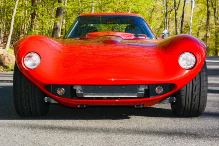 Bill Thomas Cheetah - Page 1 - General Gassing - PistonHeads - The image depicts a striking red sports car parked on a paved surface. The car's design is sleek with a low-profile stance, and its two round headlights are prominent. The car's hood is closed, and the wheels appear to be in good condition, possibly with a chrome lip. The background is a serene setting of trees partially obscured by the car's presence, suggesting the car is in a parking lot or a uncrowded roadside. The overall composition is a clear shot of the car, showcasing its design and the setting in which it is situated.