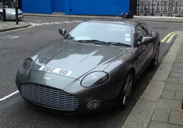 Supercars Rarities Pistonheads Spotted - The image shows a gray Aston Martin DB7 car parked on the side of a street. It is a photo taken from the side of the vehicle. The car has a single white licence plate. The street upon which the car is parked has a marked pedestrian crossing. In the background, the corner of a blue building can be seen.