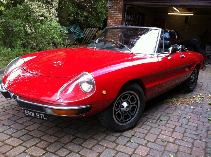 Pistonheads - The image shows a classic red sports car parked on a brick driveway. It appears to be a two-door, convertible model with a hardtop. The vehicle has chrome and black detailing, accentuating its vintage style. Behind the car, there is a garage and garden area, indicating the car is parked in a home driveway. There is no visible text on the car, and the license plate has been obscured. The setting appears to be an outdoor residential area.