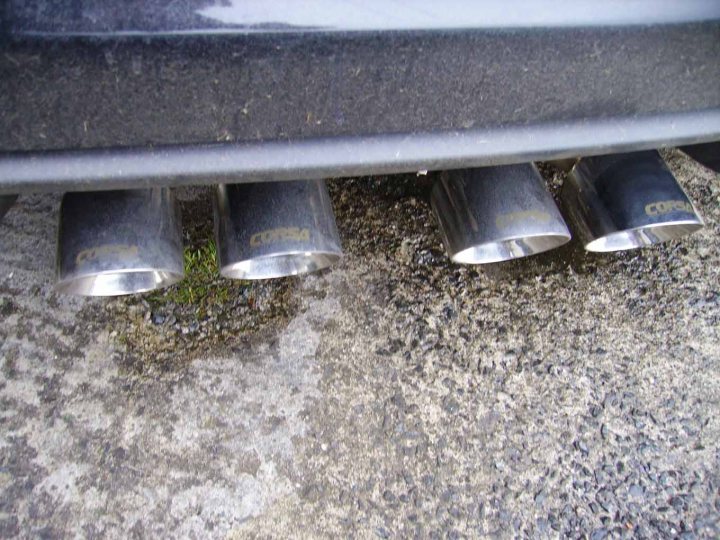 Pistonheads - The image is a close-up view of a vehicle's bumper and chrome exhaust pipes. The most prominent feature is the row of three shiny, metallic tailpipes with embossed text on each. Each of the pipes shows some text that appears to be reversed, suggesting the image is taken from an underside perspective. The rusty, uneven background suggests the image might have been taken in a parking lot or a similar functional outdoor space where the pavement is not in pristine condition. The style of the image is photographic with a focus on the vehicle components, highlighting their designs and textures.