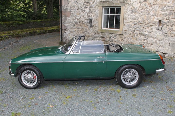 Thoughts On This £12K MGB - Page 1 - MG - PistonHeads - The image features a green vintage car, likely a Cooper S, parked on a gravel surface in front of a stone building. The car is a two-door model with a convertible top, showcasing its convertible design. The interior is sparsely visible, suggesting that the top is down and the vehicle is situated against the building's facade, under what appears to be bright sunlight. The background indicates a rural or semi-rural setting, with greenery and trees visible on either side of the scene.