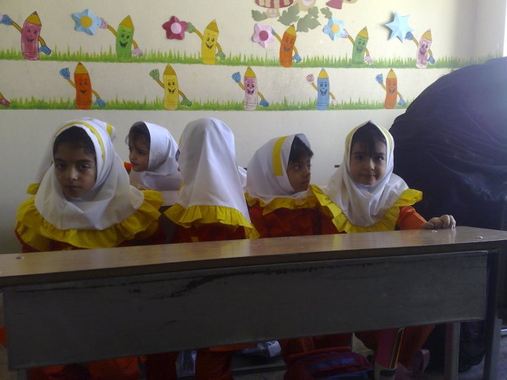 The image shows a group of children facing the camera. They appear to be in a classroom setting, with each child wearing predominantly white clothing. There are decorations on the wall behind them, including what looks like drawings and colorful posters. The children have various expressions, and some have their heads covered with white fabric. They are seated at a metal desk.