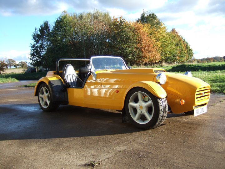 Pistonheads - The image showcases a bright yellow sports car parked on a brown road covered in fallen leaf-litter. Behind the car, multiple tall trees with autumnal foliage suggest that the photo was taken in the fall season. The vehicle's design features a convertible top and a prominent front grille, adding to its sporty appearance. The weather appears overcast with a hint of sunlight, casting a soft luminance on the scene.