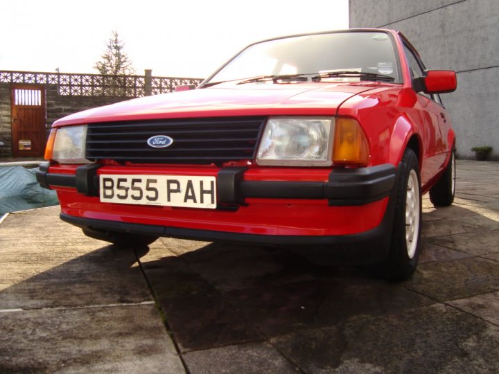 Feel like I've won the lottery - Page 1 - Classic Cars and Yesterday's Heroes - PistonHeads - The image features a vibrant red Ford Escort parked on a brick pavement. The car is of an older model, as indicated by its distinctive design, black grill, and LP badge. The vehicle is positioned at an angle, showcasing its right side, and its license plate reads "BE55 PH". In the background, there is a stone fence and foliage, suggesting a suburban or residential area. The sunlight is casting a warm glow on the car, highlighting its glossy red finish.