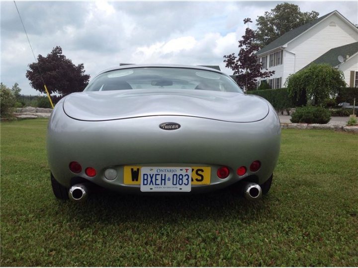 Canadians - what UK cars would you ideally like? - Page 1 - USA & Canada - PistonHeads - The image shows a silver sports car parked on a grassy area in front of a house. The car is facing the camera, and its sleek design is accentuated by its shiny silver finish. The car is equipped with two tail pipes and has a European-style license plate visible on the rear. The scene is serene, with a lawn stretching out to a driveway extending to the right. A house with a green roof can be seen in the background, partially obscured by some trees and bushes.