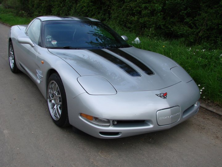 Corvette Pistonheads Hire - The image captures a sleek, gray sports car parked on what appears to be a grassy area alongside a road. The long, front view accentuates the car's low-slung design with a particularly wide scoop on the side of the vehicle, a characteristic novelty dial, and a lion badge on the grille. The circular black wheels and curved windshield create a streamlined, aerodynamic appearance. The vehicle is positioned at a slight angle to provide a clear view of its lines and features.