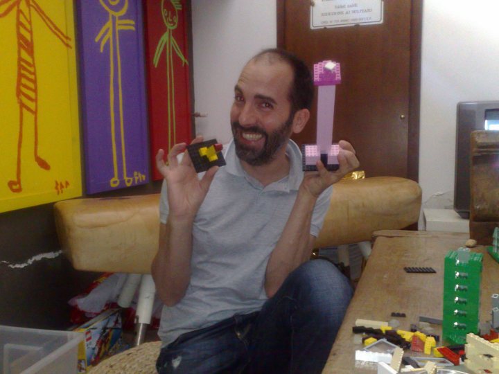 In the heart of a vibrantly colored room, a man with a warm smile on his face is proudly showing off his creations. He's holding up two miniature models - a black and yellow model and a green model, both intricately designed. The models are placed against a backdrop of various other toys and models, creating an atmosphere of creative exploration and playful craftsmanship. The man's excitement is palpable as he presents his work, whether it's a passion project or a hobby he enjoys. The image captures the essence of creativity and joy that comes from hands-on building and designing.