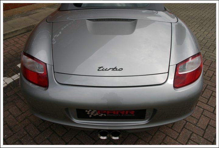 Black paint code for R spoiler, vents and mirrors - Page 1 - Boxster/Cayman - PistonHeads - The image features a sporty, silver two-door car parked on a paved surface, possibly a street or a parking lot. The car appears to be a personal vehicle, possibly owned by an individual interested in high-performance or luxury cars. The model is identifiable with a visible badge that includes the name "TURBO," and there's a Febreze logo sticker on the back of the car. A blurred logo on the front grille suggests it might be a luxury or performance brand, perhaps BMW given the context provided by the badge, which is often associated with BMW's performance models. The car is slightly angled to the left in the frame, but the details remain clear, indicating that the car is in good condition and well-maintained.