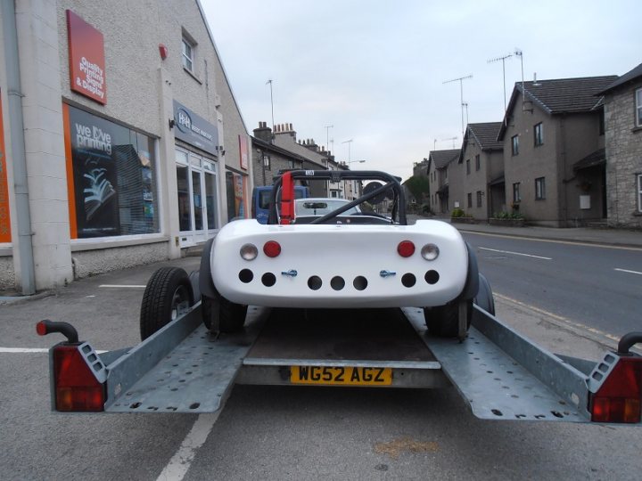 whats an ideal budget trailer for clubsport/prosport etc? - Page 1 - Radical - PistonHeads - In the image, a white vintage car is being towed on a trailer down a street lined with brick houses. The trailer, equipped with four large wheels and reflectors on its rear, is attached to a truck. The vintage car is distinct, with interesting headlights and a noticeable black grill. The truck pulling the trailer is captured in the foreground, with its cab facing the right side of the image. The scene suggests a vintage vehicle is being transported, possibly for restoration or display.