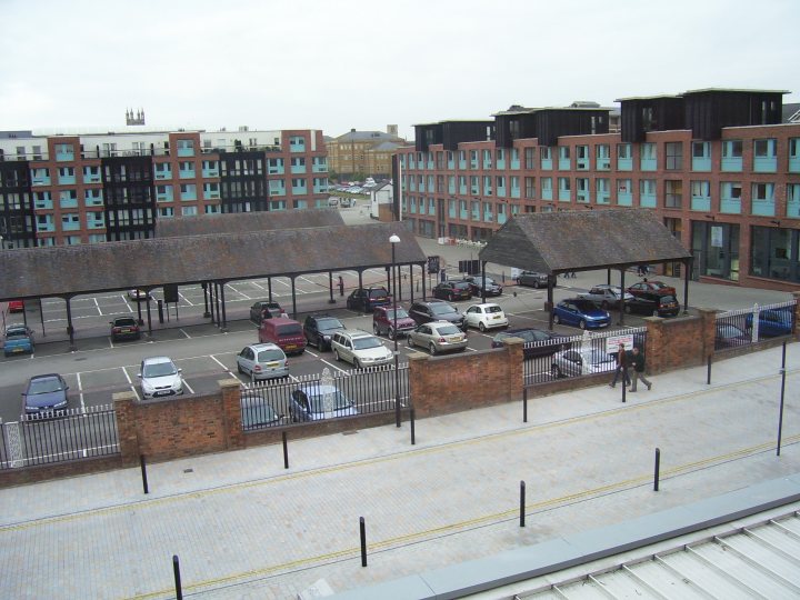 Western Pistonheads Region Owners October - The image shows an urban parking area adjacent to a collection of multi-story buildings. The parking lot contains several rows of designated parking spaces, all currently empty. A pathway runs adjacent to the parking lot, with a man walking along it. The buildings have a modern architectural style with terracotta roofs, and the overall scene suggests a quiet, possibly early morning, time of day.