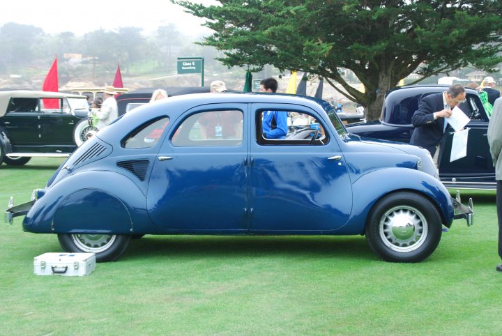 The wildest car at Pebble Beach....a Hoffman X8 - Page 1 - Classic Cars and Yesterday's Heroes - PistonHeads - The image features a classic navy blue car prominently displayed on a lush green grass lawn. The car's vintage design is complemented by a white cornet next to its front wheel, suggesting that it may be on show. The automobile is set outdoors amidst natural surroundings, marked by a path or driveway running through the background. There are multiple people in the scene, some are near the car, while others are scattered throughout the image, all dressed in casual attire, likely indicating an event or gathering where the car is the focal point. Additionally, there are some umbrellas visible in the distance, possibly suggesting an outdoor setup for an event or gathering.