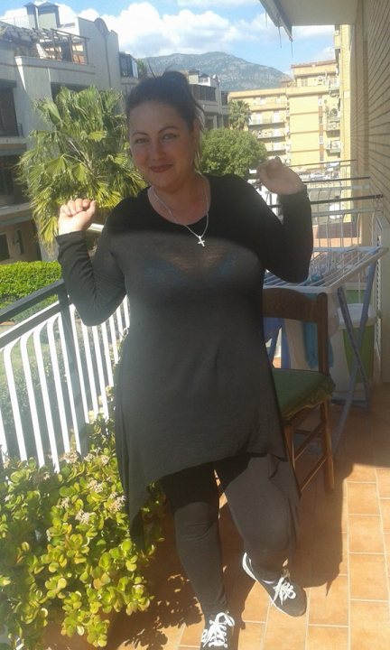 vakantie - The image features a woman standing on a balcony, posing for the camera. She is wearing a black t-shirt and has her hand on her hip. The balcony overlooks a cityscape with buildings in the background. There's also an outdoor dining table visible, suggesting that this is a residential space. The woman is smiling and appears to be enjoying the view from her balcony.