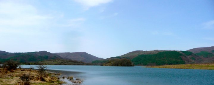 Couple of pics from today... - Page 1 - Off Road - PistonHeads - The image showcases a serene landscape dominated by a tranquil lake of dark hues. The lake stretches from the foreground towards the middle of the frame, its calm surface reflecting the surrounding hills. These hills, with their lush greenery, rise on either side of the lake, adding a sense of depth and scale to the scene. Above, a clear blue sky stretches out, its vastness underlining the isolation of the lake. A sparse scattering of trees on the left and right ends of the frame adds to the overall tranquility of the scene.