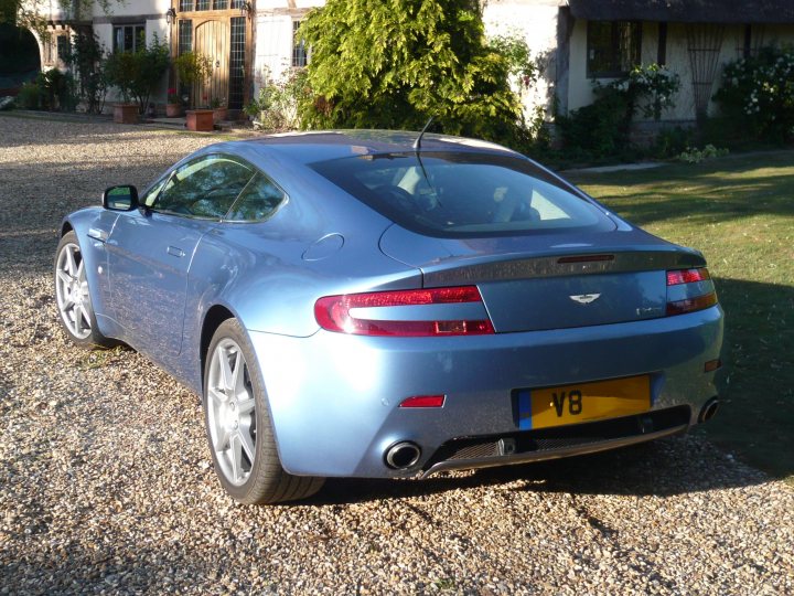 Pistonheads Ish Newbie - This image shows a blue Aston Martin car parked on a gravel drive. The car is positioned facing to the right of the frame, prominently displaying its front license plate with the letters "V8." The Aston Martin logo is visible on the back part of the car. The setting suggests a residential area, with other houses visible in the background, partially obscured by a tree. The environment has a calm and upscale feel, typical of a suburban neighborhood.